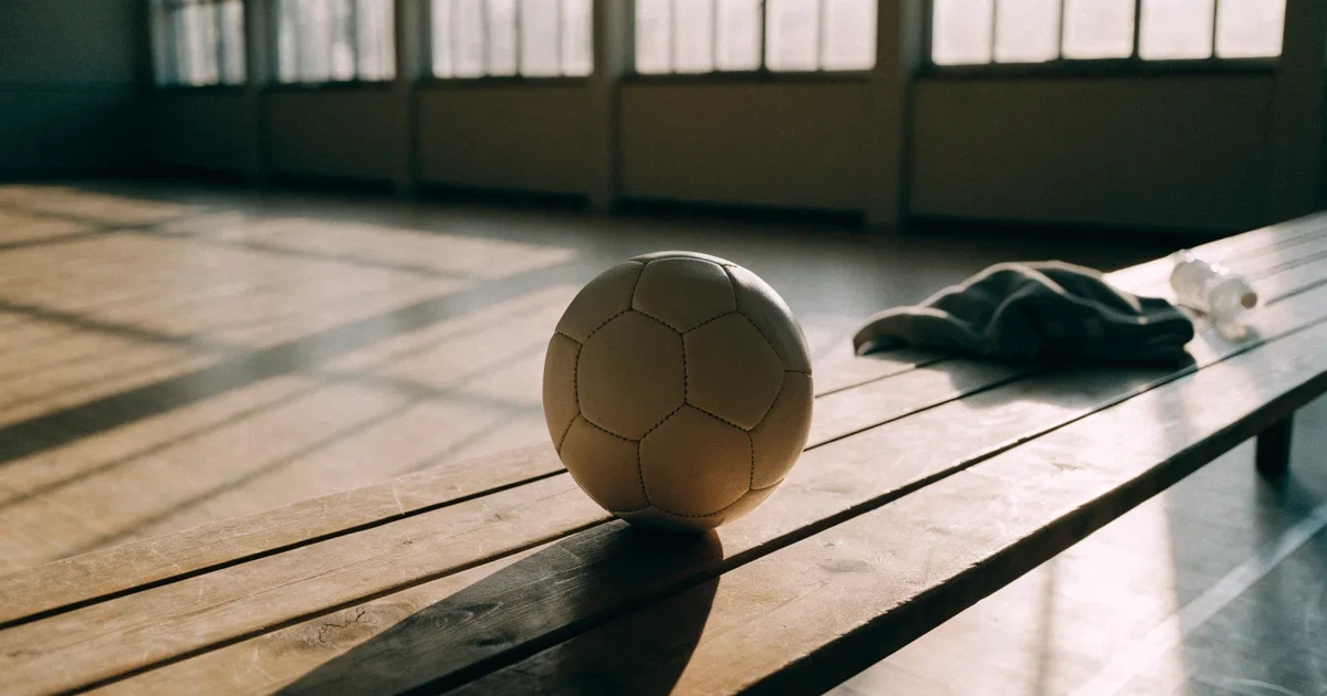 Fünf Sportbälle verschiedener Sportarten stehen in einer Reihe auf einer Holzbank in einer Turnhalle, Licht fällt durch hohe Fenster.