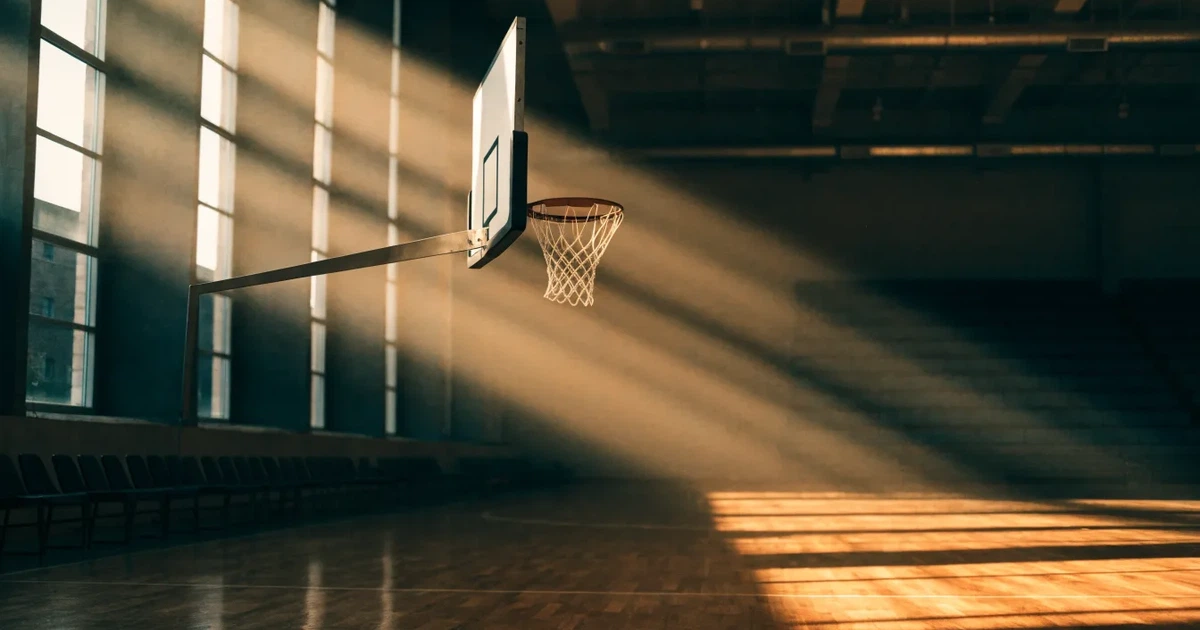 Ein Basketballkorb mit Netz in einer leeren Halle, Licht fällt von hohen Fenstern auf den glänzenden Parkettboden.